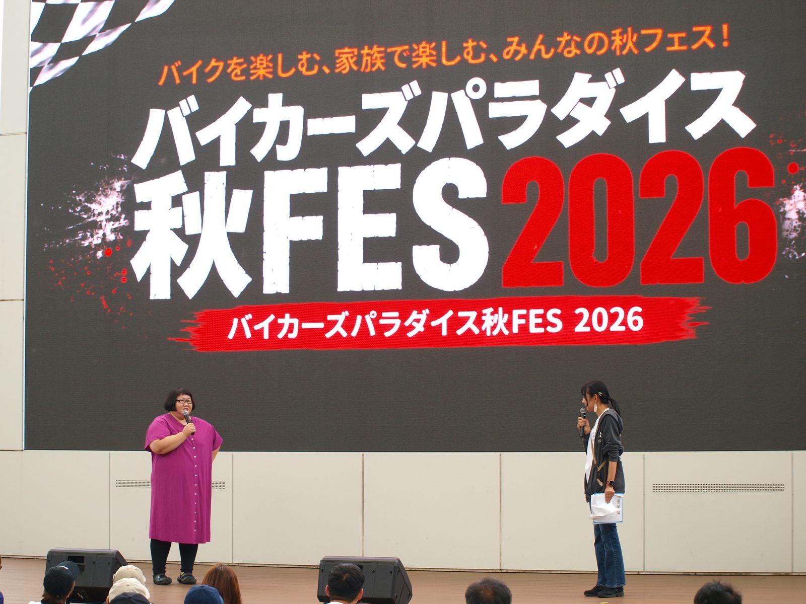 「バイカーズパラダイス秋FES 2026」の大型ビジョンの前で、ゲストとトークを進行する様子